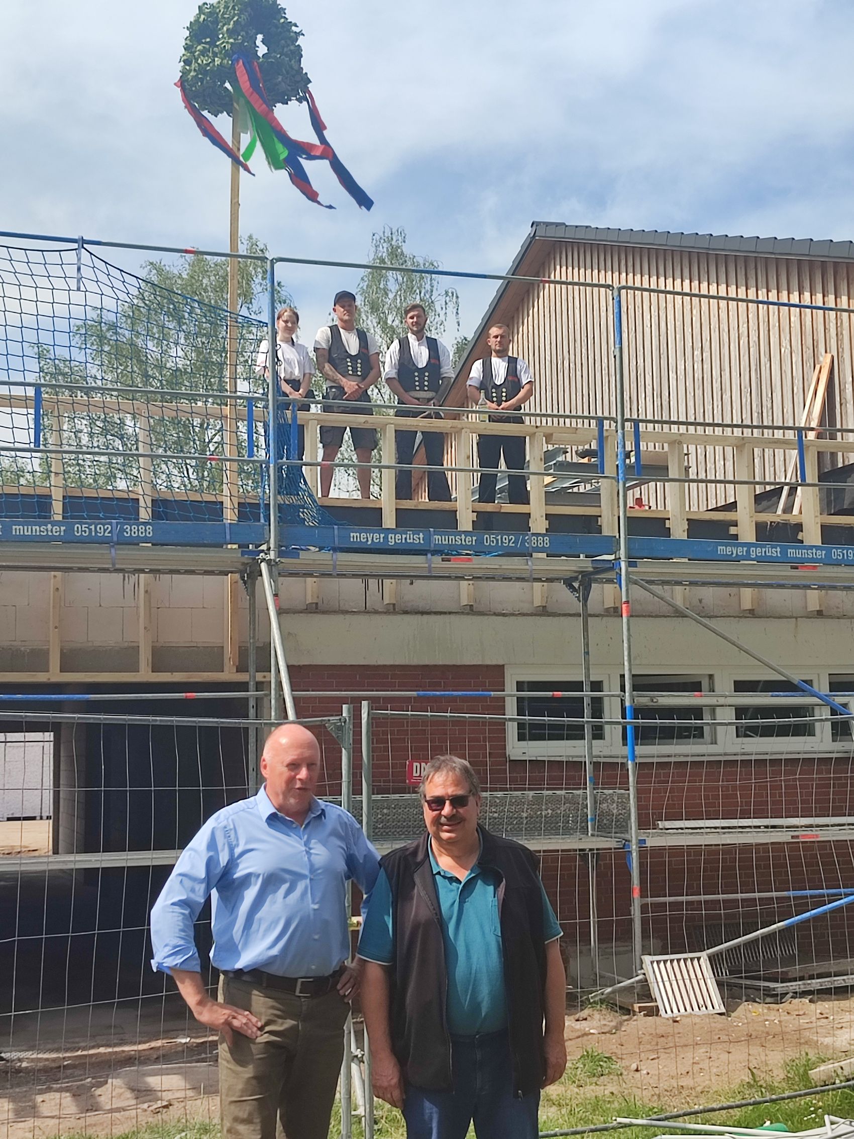 Das Bild zeigt Bürgermeister Dr. Jens Bülthuis und Detlev Loos, 1. Vorsitzender der Sportfreunde Bispingen vor der Turnhalle Hützel. Im Hintergrund stehen drei Handwerker vor der Richtkrone.