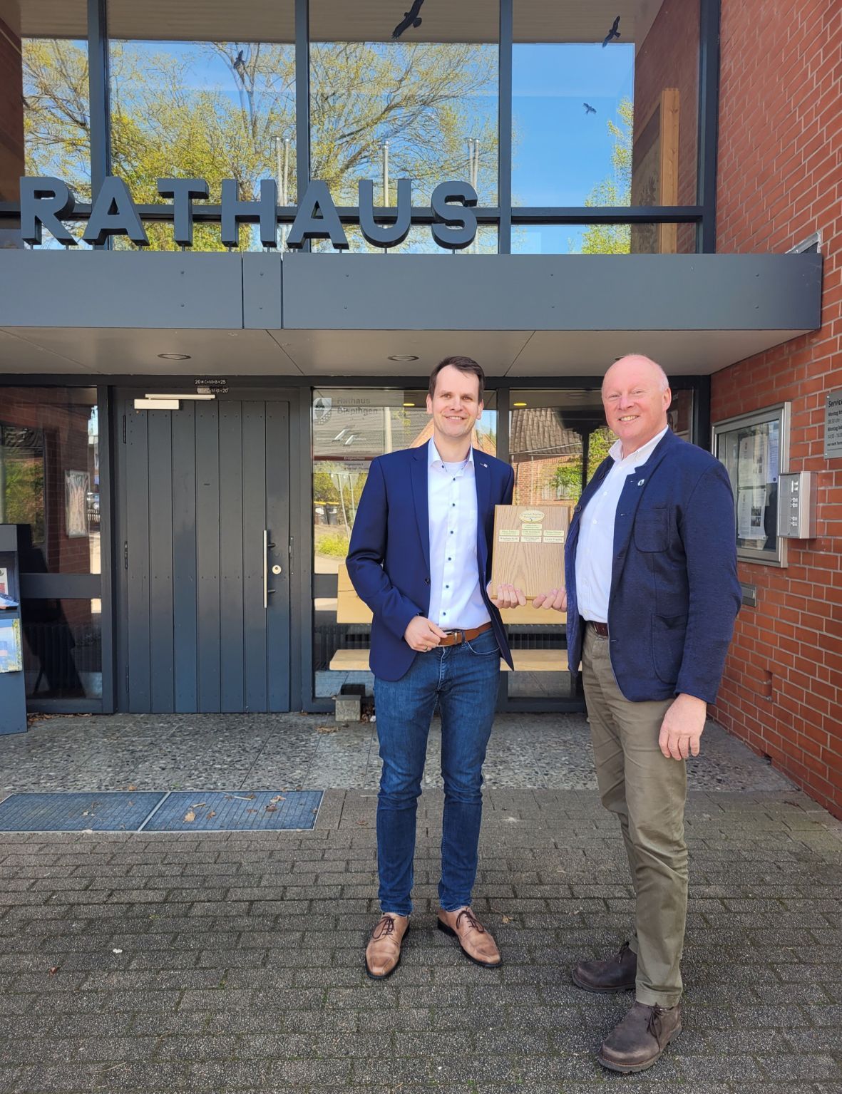 KSK-Geschäftsstellenleiter Ralf Stölpe und Bürgermeister Dr. Jens Bülthuis stehen vor dem Bispinger Rathaus. In der Hand halten sie die Tafel mit den bisherigen Preisträger*innen.