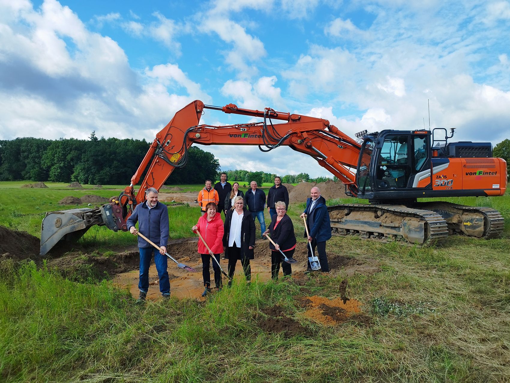 Sylvia Rose und Ulf Dallmann vom Fachbereich Planen und Bauen beim symbolischen ersten Spatenstich. Das Bild zeigt einen Bagger vor dem Rolf Scheibel (GEB), Andrea Melcher (Immobilienvermittlerin der VBLH), Laura Arp (GEB), Sylvia Rose (Gemeinde Bispingen) und Ulf Dallmann (Gemeinde Bispingen) mit Spaten stehen.
