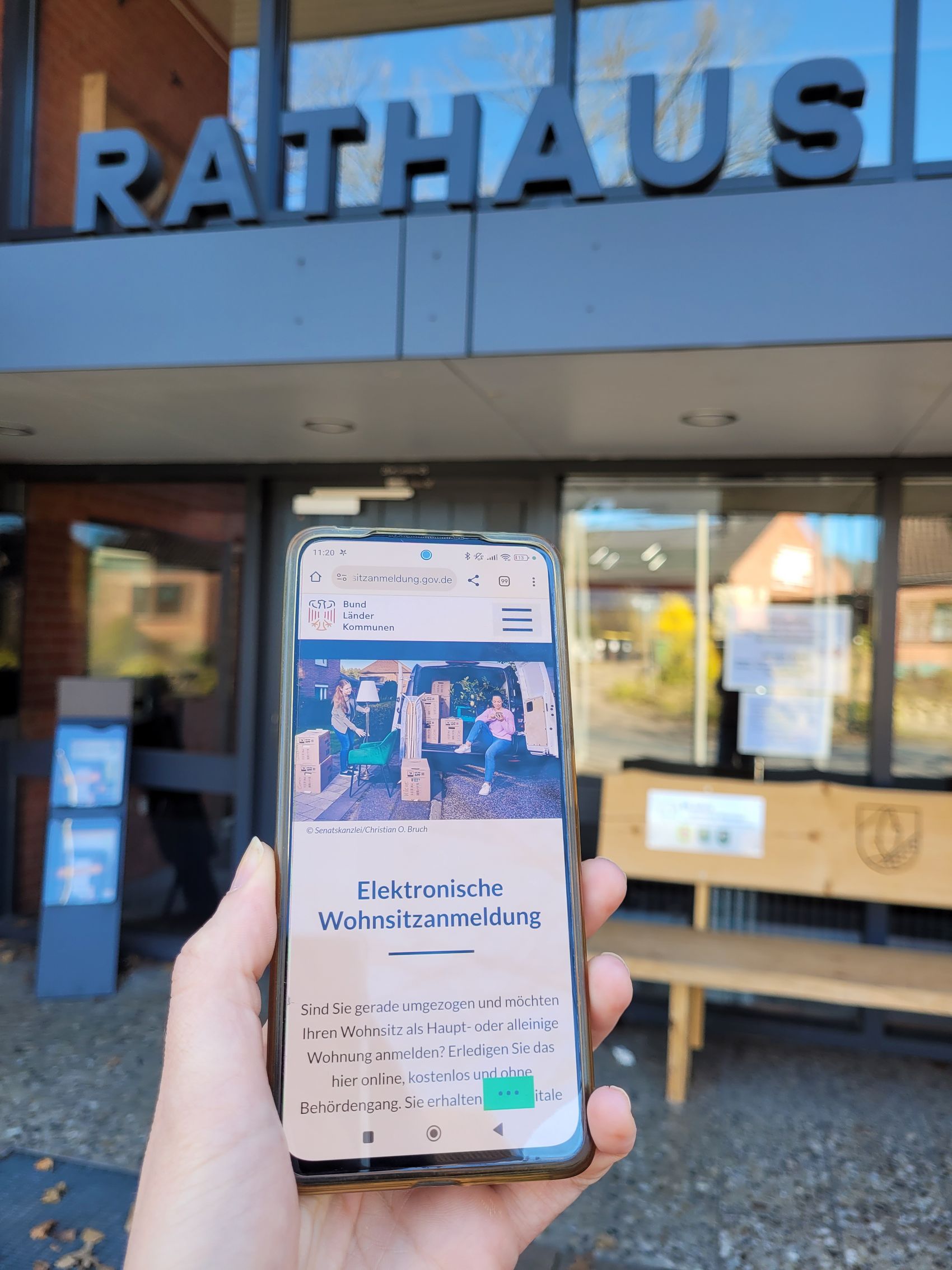 Das Bild zeigt den Eingang eines Rathauses mit großen, silbernen Buchstaben, die das Wort „RATHAUS“ über der Tür formen. Eine Person hält im Vordergrund ein Smartphone in der Hand. Auf dem Bildschirm ist eine Website mit der Überschrift „Elektronische Wohnsitzanmeldung“ zu sehen.