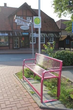 Rosa Mitfahrbank auf EDEKA-Parkplatz in Bispingen, im Hintergrund Laden 'Der kleine Speicher' und Straßenschild 'Borstel'
