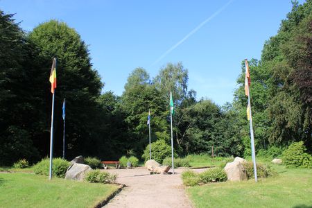 Weg im 5x Behringen Garten in Behringen mit fünf Fahnen auf Fahnenmasten, umgeben von Bäumen und Büschen