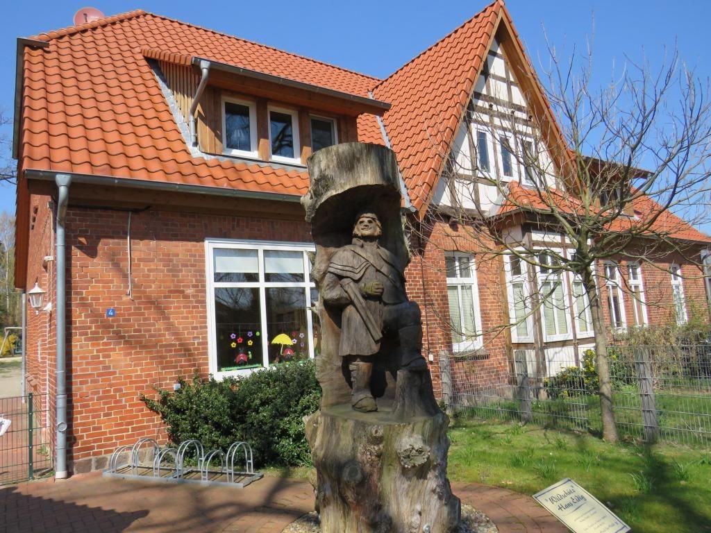Holzskulptur von Hans Eidig vor der Kita Steinbeck, zeigt eine stehende Figur mit Mantel und Hut, umgeben von einem Baumstamm