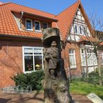 Holzskulptur von Hans Eidig vor der Kita Steinbeck, zeigt eine stehende Figur mit Mantel und Hut, umgeben von einem Baumstamm