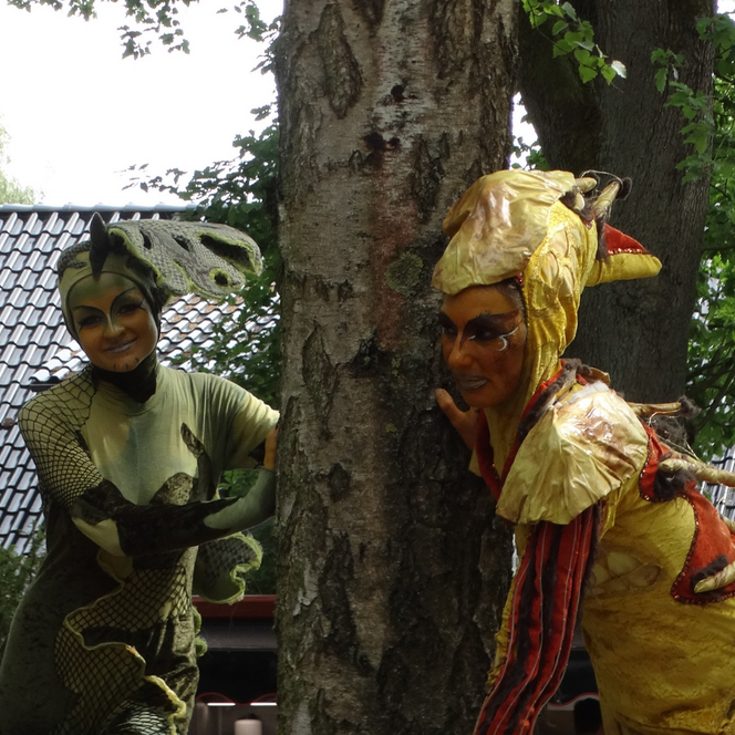 Zwei Personen in detailreichen Tierkostümen, eine als Echse mit grün-schwarzem Muster, die andere als gelb-roter Drache, stehen an einem Baum im Freien.