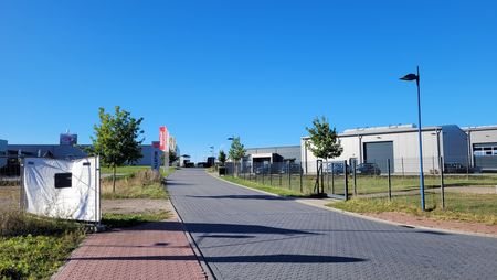 Gepflasterte Straße in einem Baugebiet an der Autobahn, rechts moderne Industriegebäude, links Bäume, blauer Himmel.