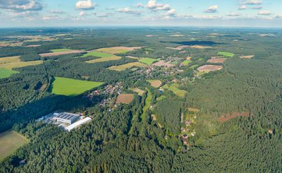 Luftbildaufnahme einer ländlichen Landschaft mit Wäldern, Feldern und verstreuten Siedlungen. Im Vordergrund sind grüne Wiesen und Bäume sichtbar.