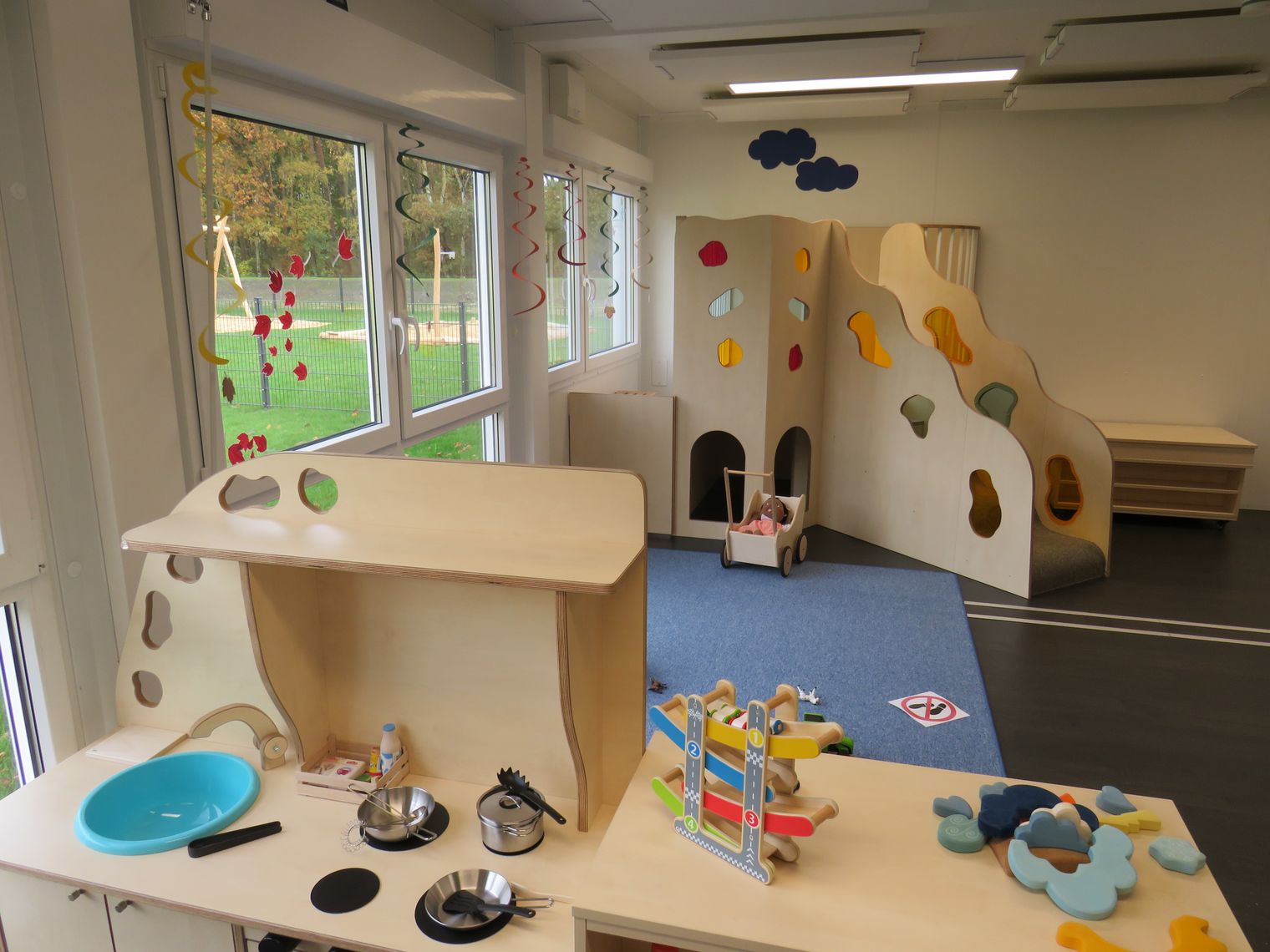 Innenansicht der Kita Wirbelwind mit Holzspielküche, Kletterwand mit bunten Griffen und verschiedenen Spielzeugen auf blauem Teppich