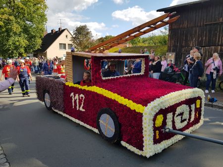 Ein mit Blumen geschmückter Wagen in Form eines roten Feuerwehrfahrzeugs mit der Nummer '112' fährt beim Erntefest Steinbeck. Im Hintergrund sind Menschen zu sehen.