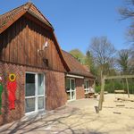 Außenansicht der Kita Sonnenblume mit rotem Backsteingebäude, Holzgiebel, zwei bunten Kinderfiguren an der Wand und Sandspielplatz mit Schaukel