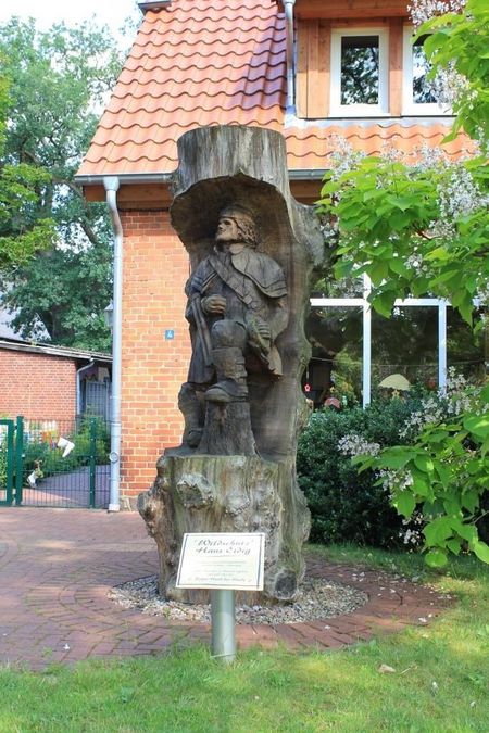 Hans Eidig Denkmal aus Holz zeigt eine stehende Figur mit Umhang und Hut vor einem Haus mit rotem Ziegeldach