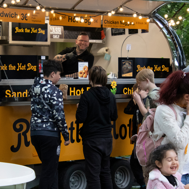 Menschen stehen vor einem gelben Imbisswagen mit der Aufschrift 'Rock the Hot Dog' und Speisekarten für Sandwich, Hot Dog und Chicken Nuggets.