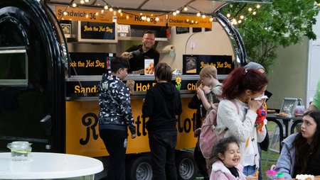 Menschen stehen vor einem gelben Imbisswagen mit der Aufschrift 'Rock the Hot Dog' und Speisekarten für Sandwich, Hot Dog und Chicken Nuggets.