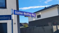 Blaues Straßenschild mit weißer Schrift 'Günther-Weißflog-Weg' an silbernem Pfosten vor Wohnhäusern und blauem Himmel.