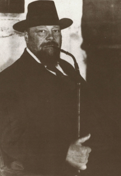 Pastor Wilhelm Bode trägt einen dunklen Anzug und einen Hut, hält eine Pfeife in der Hand, steht vor einer Wand.