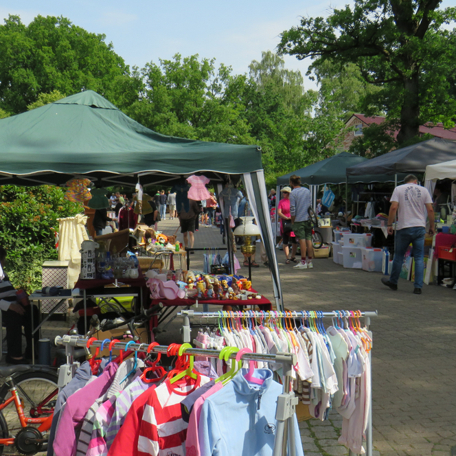 Verkaufsstände mit Zeltdächern auf einem Flohmarkt, Kleider auf Kleiderbügeln, Menschen schlendern zwischen den Ständen auf gepflastertem Weg.