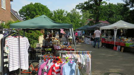 Verkaufsstände mit Zeltdächern auf einem Flohmarkt, Kleider auf Kleiderbügeln, Menschen schlendern zwischen den Ständen auf gepflastertem Weg.