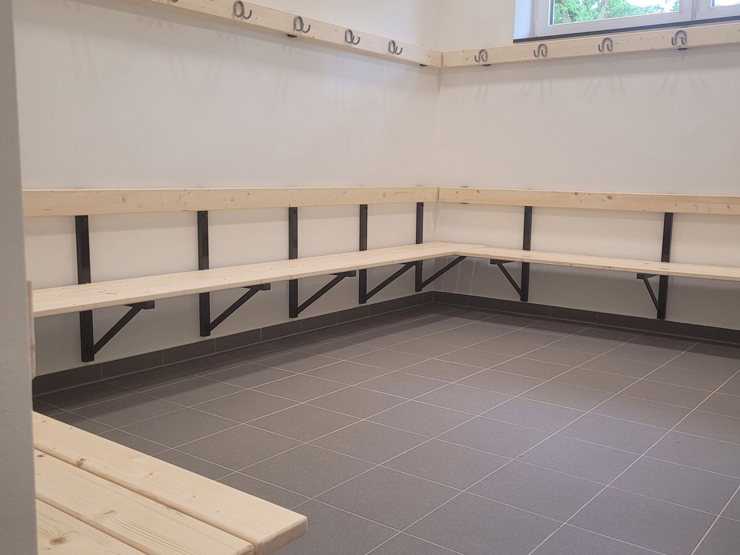 Leerer Umkleideraum mit hellen Holzbänken an zwei Wänden und Hakenleisten darüber, Fenster mit Blick ins Grüne