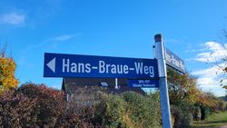 Blaues Straßenschild mit weißer Aufschrift 'Hans-Braue-Weg' und Pfeil nach links, darunter Zusatzschild mit '1895-1977 Mützeler Weidenlen' an grauem Metallpfahl, im Hintergrund Bäume und blauer Himmel.