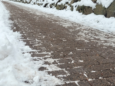 Ein gepflasterter Weg mit einer dünnen Schneeschicht ist zu sehen. Der Weg verläuft schräg im Bild und ist an einer Seite von einer niedrigen Steinmauer mit aufliegendem Schnee begrenzt. Auf dem Weg sind vereinzelt Schneereste verteilt, während die Pflastersteine größtenteils freigeräumt sind. Am Bildrand liegt eine dickere Schneeschicht neben dem Weg.
