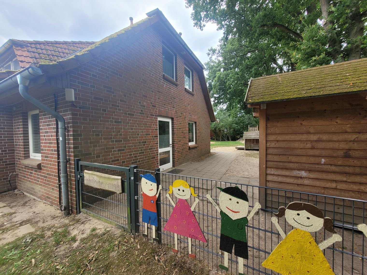 Eingangstor der Kita Hörpel mit bemalten Holzfiguren von vier Kindern am Zaun, Backsteingebäude links und Holzhütte rechts