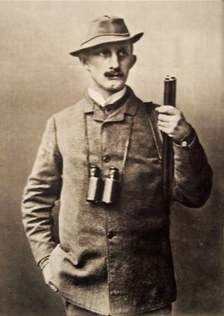 Hermann Löns um 1900 stehend mit Hut, Jagdbekleidung, Fernglas um den Hals und einem Gewehr in der rechten Hand.