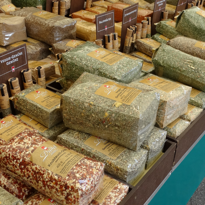 Verpackte getrocknete Kräuter und Gewürze in rechteckigen transparenten Beuteln auf einem Marktstand, beschriftete Holzschilder mit Produktnamen.