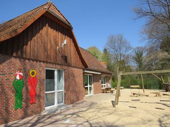 Außenansicht der Kita Sonnenblume mit rotem Backsteingebäude, Holzgiebel, zwei bunten Kinderfiguren an der Wand und Sandspielplatz mit Schaukel
