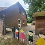 Eingangstor der Kita Hörpel mit bemalten Holzfiguren von vier Kindern am Zaun, Backsteingebäude links und Holzhütte rechts