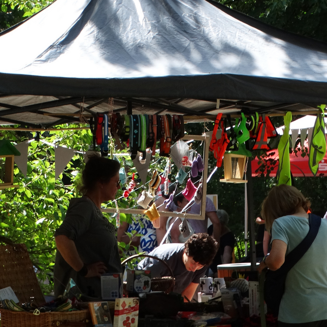 Marktstand mit Zelt, an dem bunte Hundehalsbänder und -geschirre hängen, mehrere Personen stehen davor und schauen sich die Ware an.