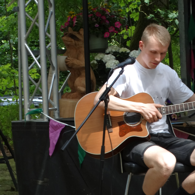 Junger Mann sitzt auf einem Stuhl, spielt akustische Gitarre, trägt weißes T-Shirt und schwarze Shorts, im Hintergrund Pflanzen und Bühnenausstattung.