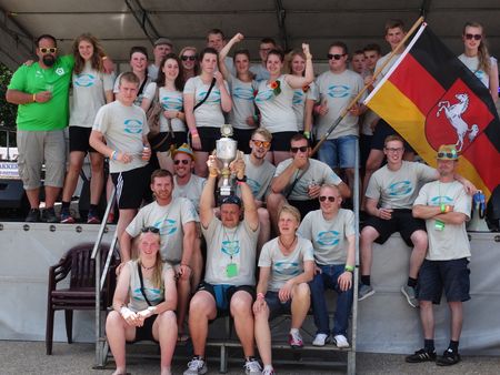 Gruppe von Personen in hellen T-Shirts mit blauem Logo, einige sitzen, andere stehen auf einer Bühne. Eine Person hält eine Fahne mit dem Niedersachsen-Wappen, eine weitere einen Pokal.