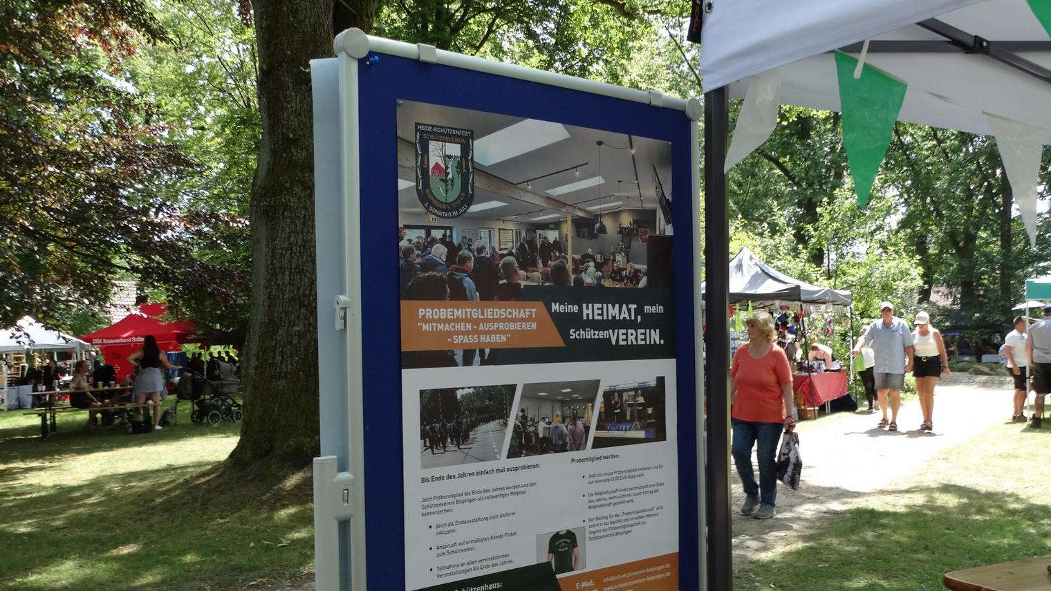 Informationsstand mit Plakat zur Probemitgliedschaft des Schützenvereins, umgeben von Bäumen und Marktständen, Menschen gehen vorbei.