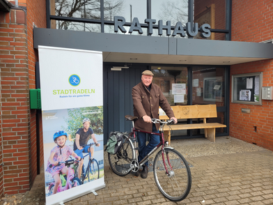Bürgermeister Dr. Jens Bülthuis steht mit einem Fahrrad vor dem Eingang des Bispinger Rathauses. Neben ihm befindet sich ein aufgestelltes Roll-up-Banner mit dem Logo und Schriftzug von Stadtradeln sowie einem Foto von zwei Radfahrenden. Die Fassade besteht aus roten Ziegeln, über dem Eingang ist in großen Buchstaben das Wort 'Rathaus' angebracht. Im Hintergrund steht eine Holzbank.