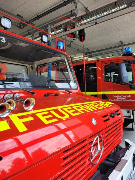 Zwei rote Feuerwehrautos der Feuerwehr Hörpel in einer Fahrzeughalle, vorne ein Mercedes mit gelber Aufschrift 'FEUERWEHR' auf der Motorhaube.