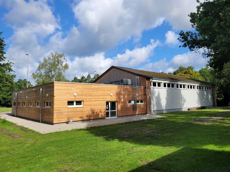 Sporthalle in Hützel mit Holzfassade und angrenzendem weißem Hallenbereich unter bewölktem Himmel