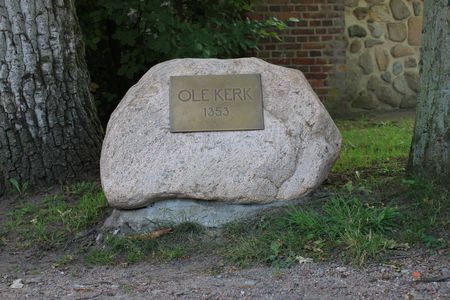 Großer Stein mit einer Metalltafel, auf der 'OLE KERK 1353' steht, zwischen zwei Bäumen vor einer Backsteinmauer.