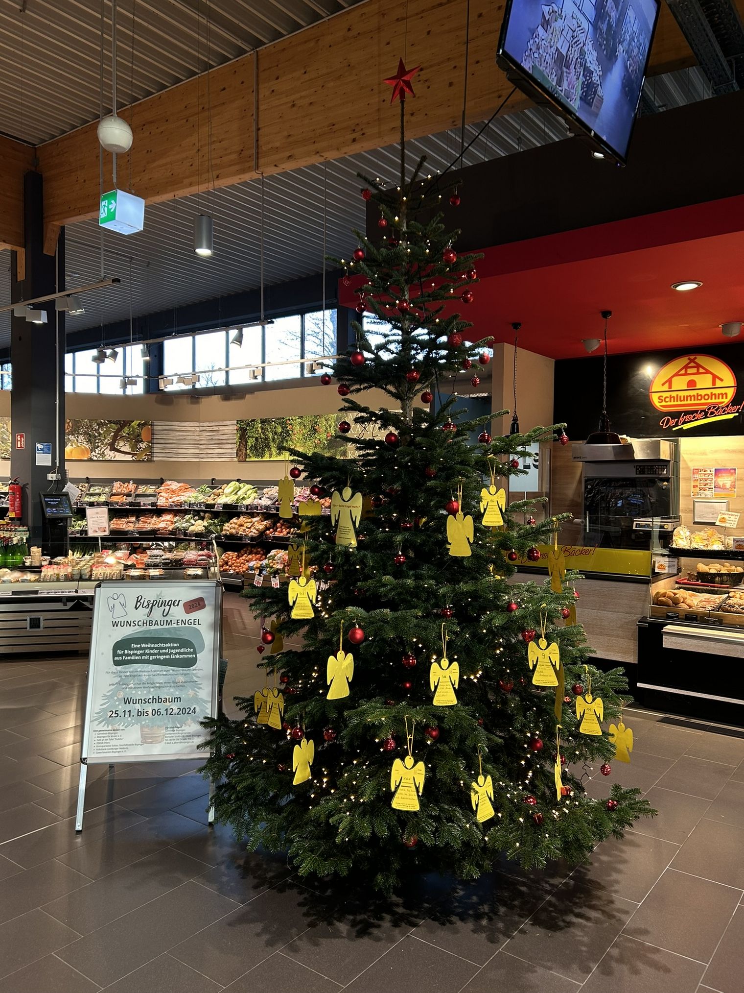 Weihnachtsbaum mit roten Kugeln und gelben Engelanhängern in einem Supermarkt, daneben ein Schild mit Informationen zur Wunschbaum-Engel Aktion.