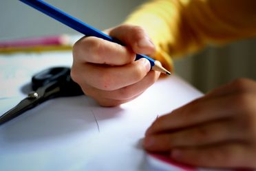 Kind schreibt mit blauem Buntstift auf weißem Papier, Schere liegt daneben