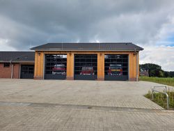 Feuerwehrhaus Hörpel mit drei großen Toren, hinter denen zwei rote Feuerwehrautos sichtbar sind, unter bewölktem Himmel.