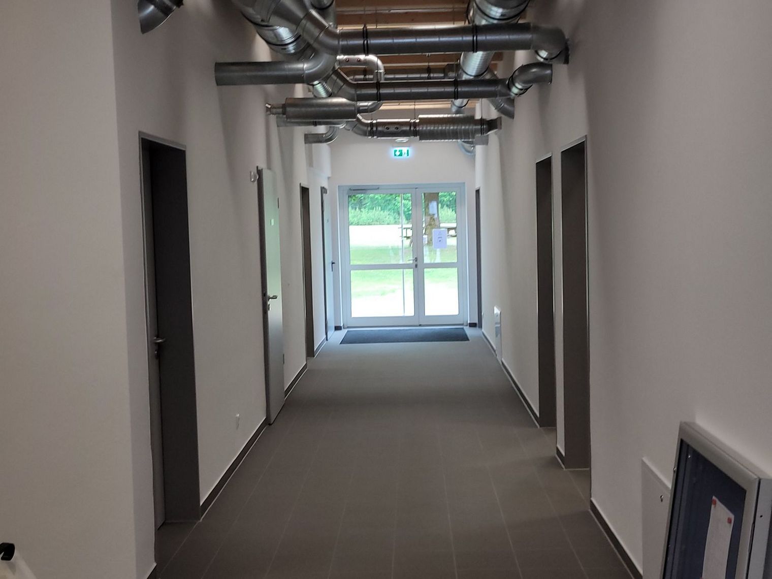 Flur mit grauem Boden und weißen Wänden, an der Decke sichtbare Lüftungsrohre, mehrere geschlossene Türen, Ausgangstür mit Blick auf grünes Außengelände