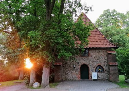 Ole Kerk Bispingen, kleine steinerne Kirche mit spitzem Ziegeldach, umgeben von Bäumen bei Sonnenuntergang
