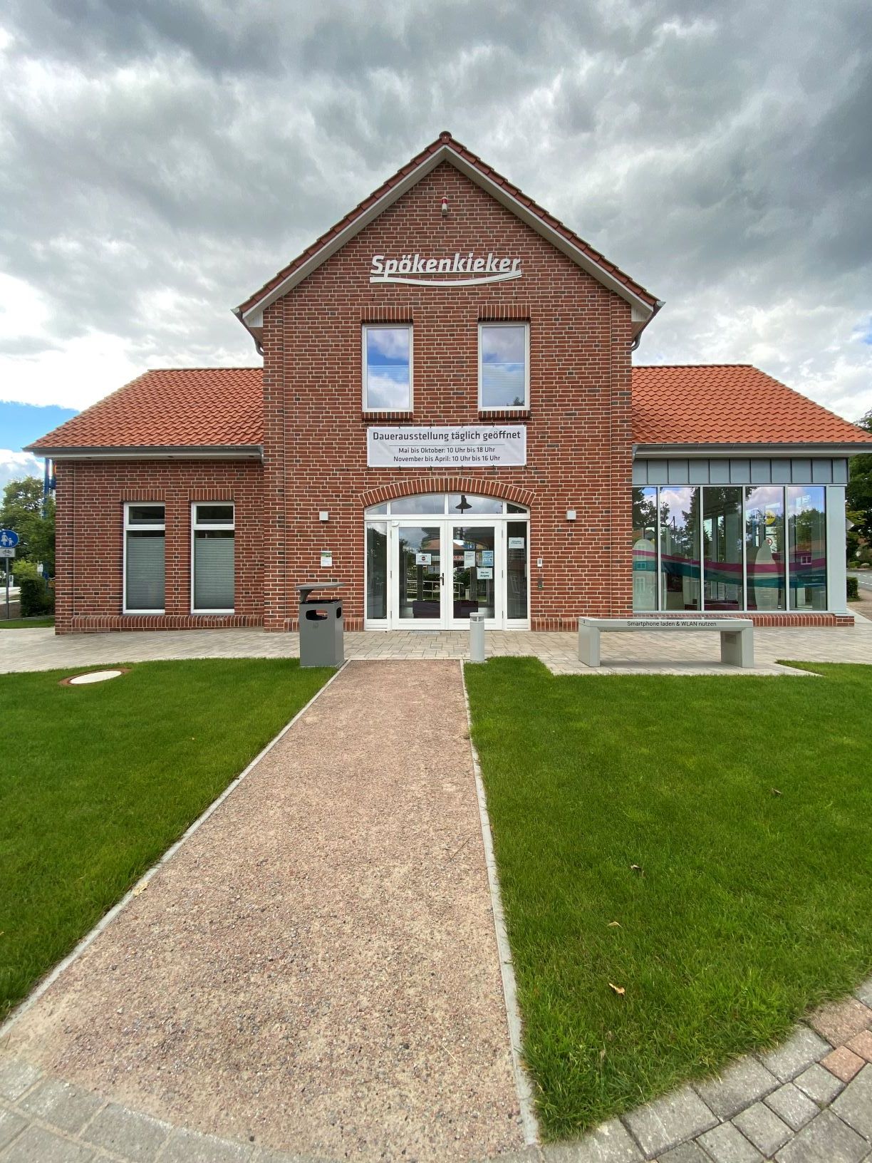 Das Bild zeigt ein einstöckiges Gebäude aus roten Ziegelsteinen mit einem zentralen, höher gebauten Giebel. Das Dach ist mit roten Ziegeln gedeckt. Vor dem Gebäude verläuft ein schmaler, gepflasterter Gehweg, der direkt zu einem weißen Doppeltür-Eingang führt. Links und rechts vom Gehweg ist eine Rasenfläche zu sehen. Das Gebäude hat große Fenster, die viel Licht hineinlassen. Über dem Eingang befinden sich zwei kleinere Fenster. Der Himmel im Hintergrund ist größtenteils bewölkt, mit einigen blauen Stellen, durch die der Himmel sichtbar ist. Die Perspektive der Aufnahme ist frontal und leicht nach unten gewölbt, was dem Bild einen leichten Fischaugeneffekt verleiht.
