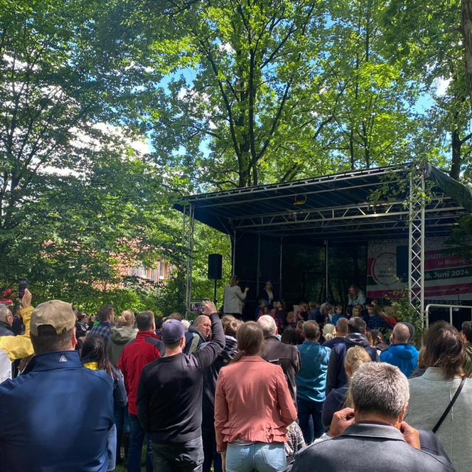 Menschenmenge vor einer kleinen Bühne im Freien, umgeben von Bäumen, bei Tageslicht auf dem Heidemarkt Bispingen.