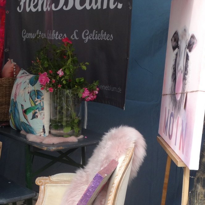 Stuhl mit rosa Fellüberzug vor Staffelei mit Hundeporträt, daneben bunte Gemälde und Blumen in Vase.