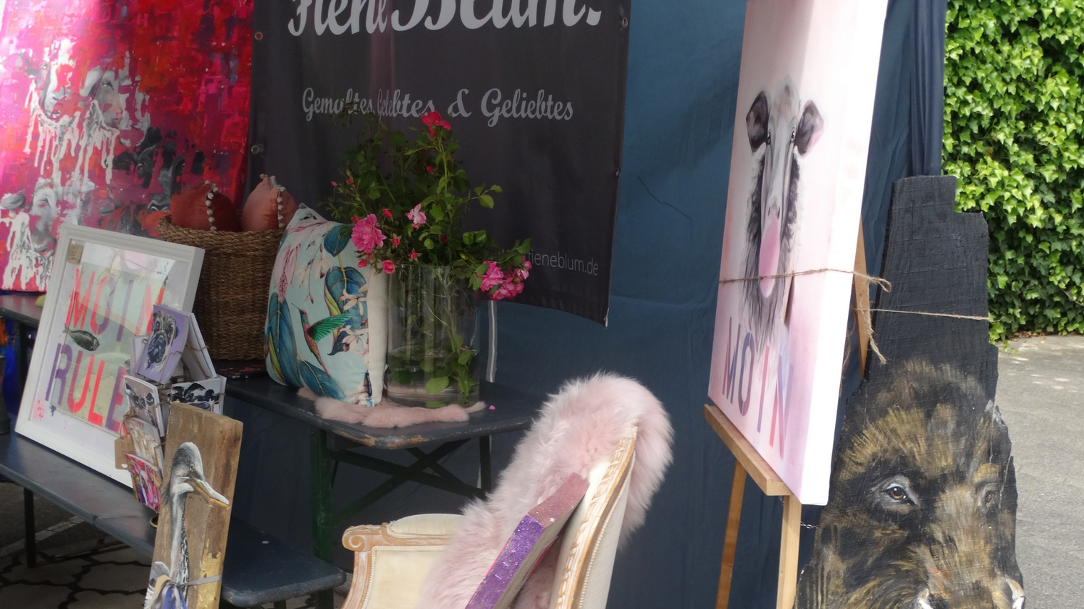 Stuhl mit rosa Fellüberzug vor Staffelei mit Hundeporträt, daneben bunte Gemälde und Blumen in Vase.