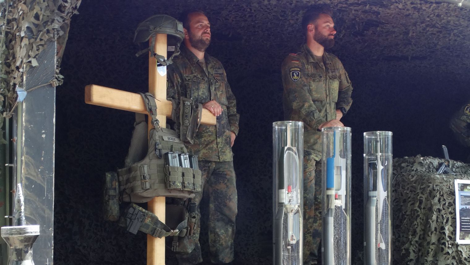 Zwei Soldaten in Tarnuniform stehen in einem mit Tarnnetz bedeckten Stand der Patenkompanie am Bispinger Heidemarkt mit Ausrüstung.