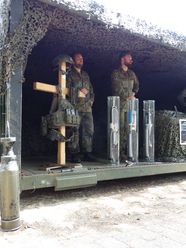 Zwei Soldaten in Tarnuniform stehen in einem mit Tarnnetz bedeckten Stand der Patenkompanie am Bispinger Heidemarkt mit Ausrüstung.