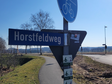 Straßenschild mit der Aufschrift 'Horstfeldweg' an einem Rad- und Fußweg unter blauem Himmel