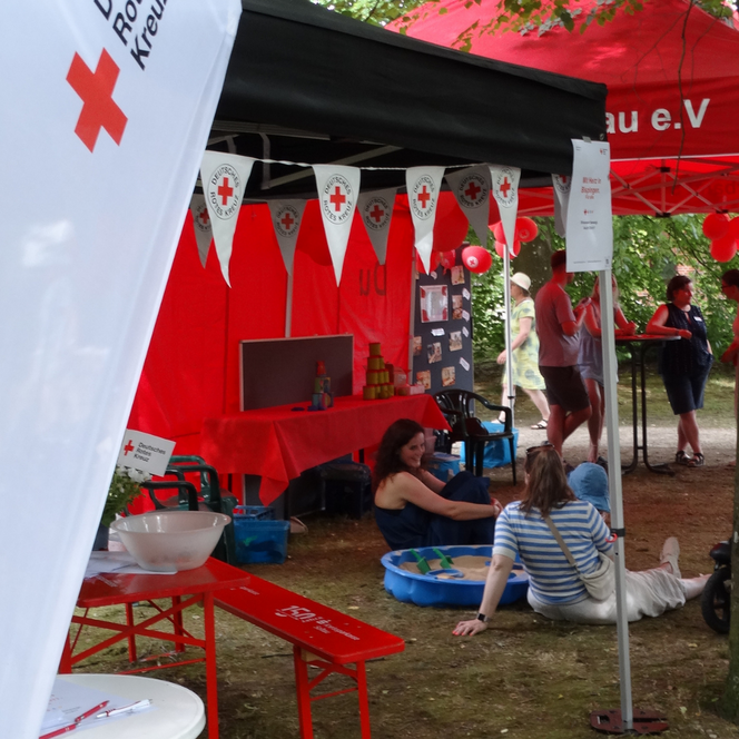 Rotes Kreuz Informationsstand mit Fahnen, Bannern und Sitzgelegenheiten im Freien, Menschen sitzen und stehen im Bereich.